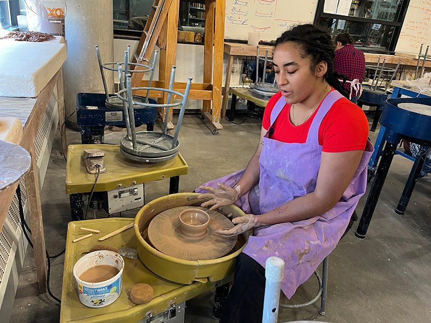在陶艺教室里，一个学生坐在陶艺轮前扔着一个小碗，背景是更多的陶艺轮.